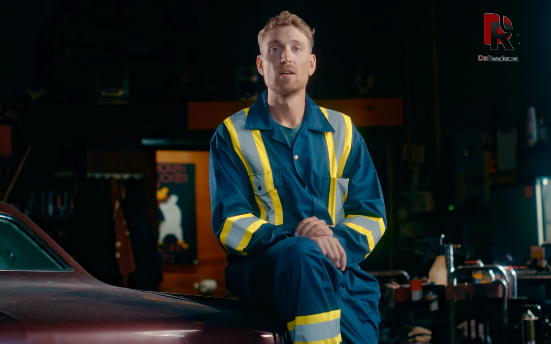 A DeReader campaign presenter seated in a cinematic workshop setting.